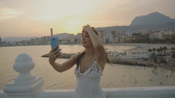 Vonzó fiatal női figyelembe selfie a smartphone. Kaukázusi nő, ami önarckép fénykép vagy video telefon megérintette a haját, a summer resort city hegyi és tengeri táj háttér. — Stock videók