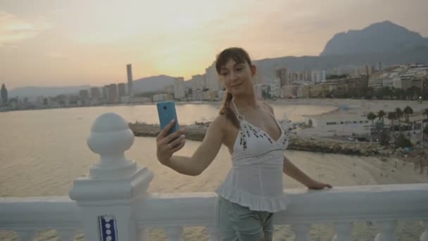 Vonzó fiatal női figyelembe selfie a smartphone. Szexi kaukázusi nő, ami önarckép fénykép vagy videó a mobiltelefon-nyári sunset resort city hegyi és tengeri táj háttér. — Stock videók