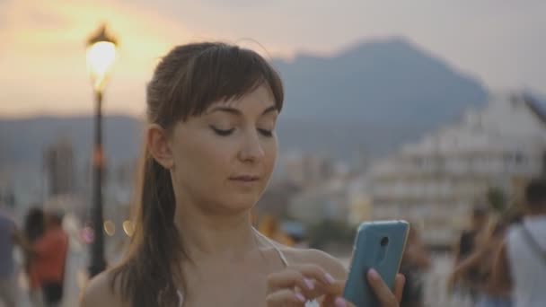 Atractiva joven hembra con smartphone. Sonriente mujer caucásica charlando en el teléfono celular en verano puesta del sol resort ciudad montaña fondo . — Vídeos de Stock