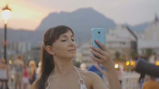 Vonzó fiatal női figyelembe selfie a smartphone. Kaukázusi nő, ami önarckép fénykép vagy video telefon megérintette a haját, a naplemente mountain resort city háttér. — Stock videók