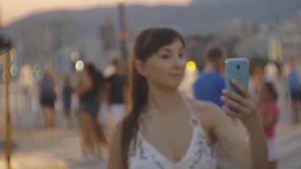 Atractiva joven hembra tomando selfie con smartphone. Mujer caucásica haciendo foto o video autorretrato con teléfono en el fondo de la ciudad del resort de montaña al atardecer . — Vídeos de Stock