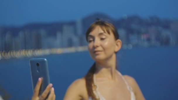 스마트폰으로 매력적인 젊은 여성 복용 selfie입니다. 섹시 한 백인 여자 여름 밤 리조트 도시 산과 바다 풍경 배경에 자기 초상화 사진이 나 휴대 전화 비디오를 만드는. — 비디오