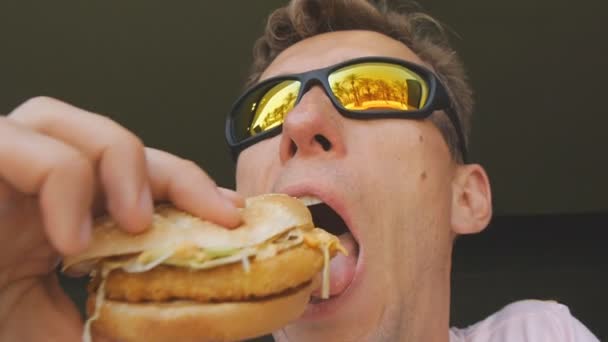 Um homem a comer comida rápida. Close-up tiro de homem caucasiano em espelho óculos de sol refletindo palmeira praia mordendo e mastigar hambúrguer no restaurante fast food . — Vídeo de Stock