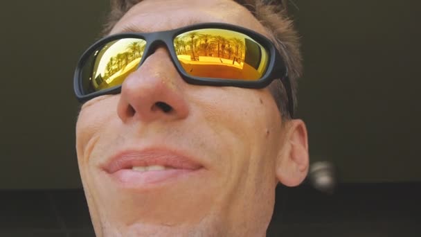 Крупним планом знімок щасливого усміхненого кавказького чоловіка в дзеркальних сонцезахисних окулярах, що відображають пляж пальмового дерева, тремтячи головою в часі з музикою. Повільний рух . — стокове відео