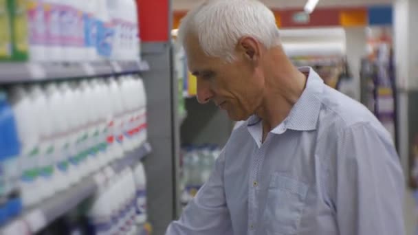 Senior man in supermarkt. Oudere grijs-haired man in blauw shirt kiezen huishoudelijke chemicaliën op opslagplanken. — Stockvideo