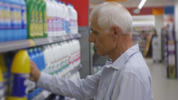 Seniorchef im Supermarkt. Älterer grauhaariger Mann in blauem Hemd wählt Haushaltschemikalien in den Regalen der Geschäfte. — Stockvideo