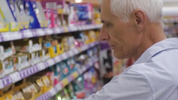 Senior man in supermarkt. Close-up van oudere grijs-haired man in blauw shirt kiezen van voedsel voor dieren op de winkel rekken achtergrond. — Stockvideo