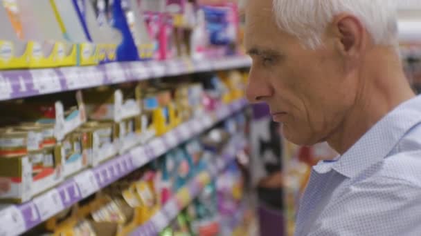 Hombre mayor en el supermercado. Primer plano del anciano hombre de pelo gris con camisa azul recogiendo lata de comida para animales en los estantes de la tienda fondo . — Vídeos de Stock