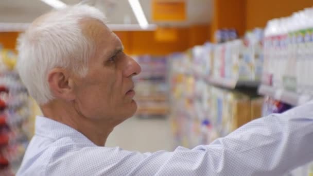 Seniorchef im Supermarkt. Nahaufnahme eines älteren grauhaarigen Mannes in blauem Hemd, der sich für Milch mit langem Leben in den Regalen entscheidet. — Stockvideo