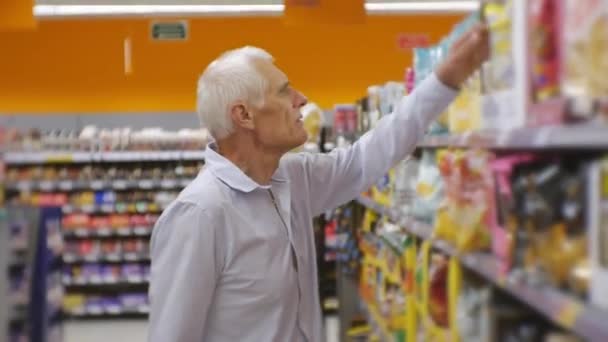 Hombre mayor en el supermercado. Anciano hombre de pelo gris en camisa azul elegir bocadillos y poner en la cesta de alimentos en los estantes de la tienda de fondo . — Vídeos de Stock