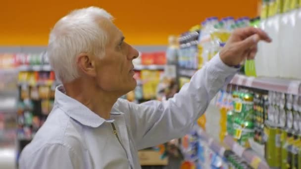 Senior au supermarché. Homme âgé aux cheveux gris en chemise bleue en choisissant des boissons gazeuses sur les étagères du magasin fond . — Video