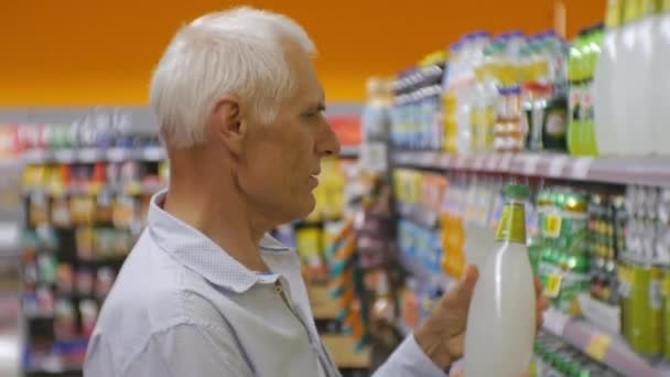 Äldre mannen i snabbköpet. Äldre gråhårige mannen i blå skjorta att välja läsk på store hyllor bakgrund. — Stockvideo