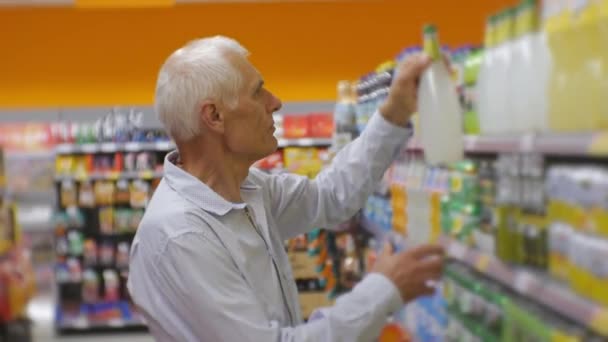 Äldre mannen i snabbköpet. Äldre gråhårige mannen i blå skjorta att välja och sätta i mat korg läsk på store hyllor bakgrund. — Stockvideo