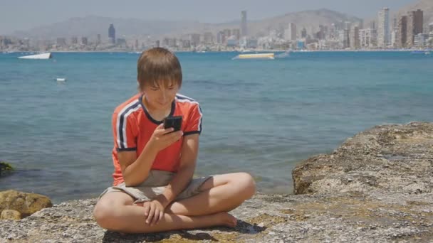 Caucasien adolescent garçon utilise smartphone regardant l'écran assis sur une jetée par la mer sur les gratte-ciel fond skyline à la journée . — Video