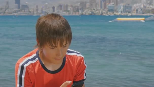 Close-up van knappe tiener jongen gebruikend smartphone kijken naar het scherm zat op een pier door de zee op de achtergrond van de skyline van wolkenkrabbers overdag. — Stockvideo