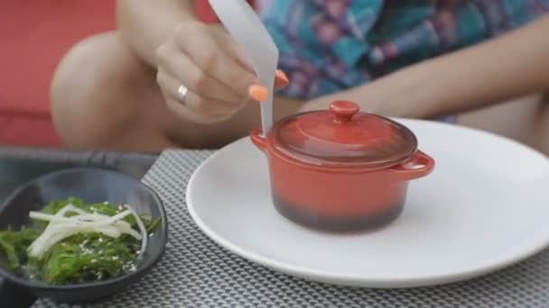 Atractiva joven caucásica abre una sartén de barro y prueba un olor a sopa en la terraza abierta del restaurante japonés . — Vídeo de stock