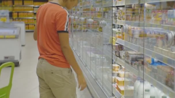 Adolescente en el supermercado. Caucásico adolescente chico en rojo camiseta elegir la leche fresca de la nevera en los estantes de la tienda fondo . — Vídeos de Stock