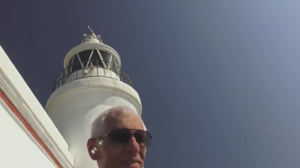 Turista jubilado anciano. Primer plano del hombre mayor de pelo gris caucásico activo en gafas de sol sobre faro blanco y fondo de cielo azul . — Vídeos de Stock