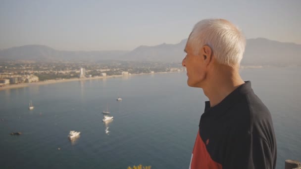 Turista jubilado anciano. Hombre mayor de pelo gris caucásico activo mira alrededor de pie en la montaña en el fondo de la costa del mar . — Vídeos de Stock