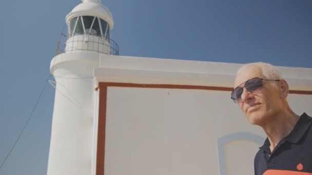 Homem idoso aposentado turista. Homem idoso caucasiano ativo de cabelos grisalhos em óculos de sol no farol branco e fundo azul do céu . — Vídeo de Stock