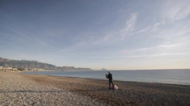 Senior man paints a picture on the beach. Wide shot of elderly male artist sets the picture on the easel on the empty sea morning beach.