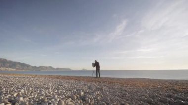 Senior man paints a picture on the beach. Wide shot of elderly male artist sets the picture on the easel on the empty sea morning beach.