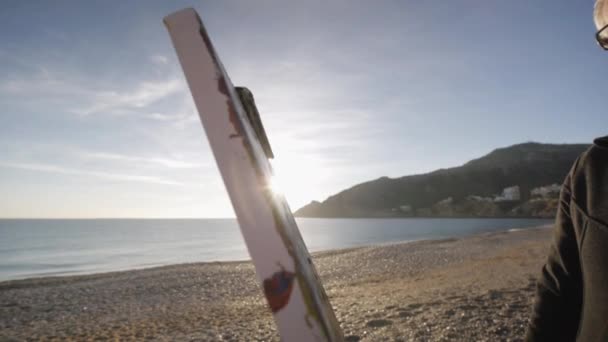 Un hombre mayor pinta un cuadro en la playa. Primer plano de la pintura de lienzo contra la elevación del sol sobre la orilla del mar y las montañas. Sol de la mañana reflejado en el tranquilo agua del mar . — Vídeo de stock