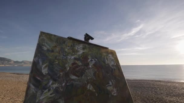 L'anziano dipinge un quadro sulla spiaggia. Vista posteriore di anziano artista maschile guardando il suo quadro dipinto di recente su sfondo spiaggia di ghiaia mare. Rallentatore . — Video Stock