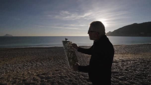 L'anziano dipinge un quadro sulla spiaggia. Anziani pitture maschili in piedi contro il sorgere del sole sulla riva del mare e sulle montagne. Sole del mattino riflesso nella calma acqua di mare . — Video Stock