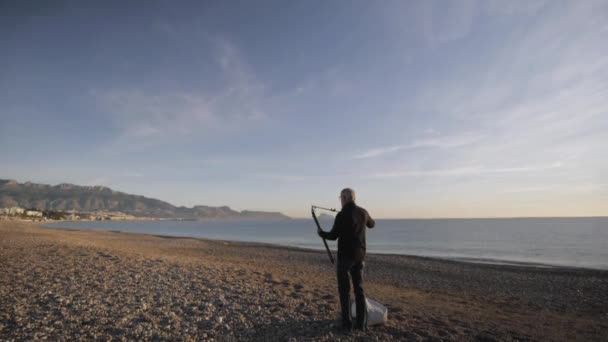 先輩の男が浜辺に絵を描く。高齢の男性アーティストは、小石の海の朝のビーチにイーゼルを設定します. — ストック動画