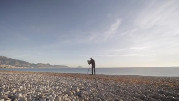 Старший чоловік малює картину на пляжі. Широкий знімок літнього чоловіка художник встановлює зображення на мольберті на порожньому морському ранковому пляжі . — стокове відео