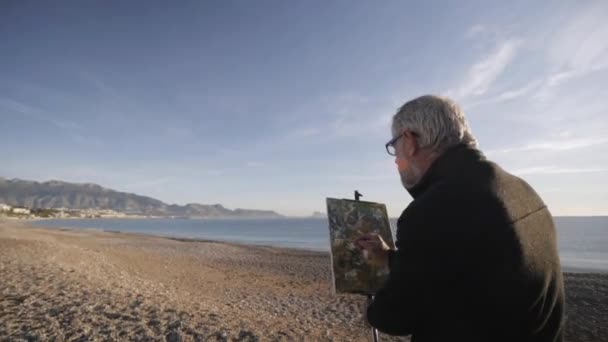 Ein älterer Mann malt ein Bild am Strand. Ein älterer männlicher Künstler gibt der modernen abstrakten Malerei gegen die aufgehende Sonne über dem Meer den letzten Schliff. Morgensonne spiegelt sich im ruhigen Meerwasser — Stockvideo