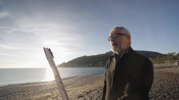 Старший чоловік малює картину на пляжі. Вид ззаду на літнього художника-чоловіка, який дивиться на його сучасну абстрактну картину проти підняття сонця над морем. Ранкове сонце відбивається у спокійній морській воді . — стокове відео