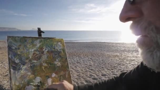 老人在海滩上画了一幅画。年长的男性艺术家在现代抽象画上用铲子快速完成润色，反对在海边升起太阳. — 图库视频影像