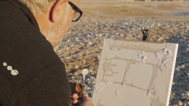 O senhor pinta um quadro na praia. Visão traseira do artista masculino idoso aplicando tinta na tela com uma espátula na praia do mar pebble sunrise . — Vídeo de Stock