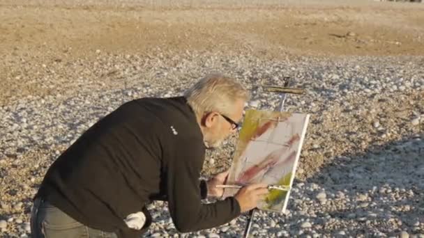 Ein älterer Mann malt ein Bild am Strand. ältere männliche Künstler malen abstraktes Bild mit einem Pinsel am Kieselstrand Sonnenaufgang Meer. — Stockvideo