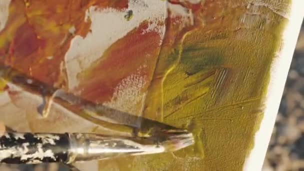 Senior man paints a picture on the beach. Close-up of elderly male artists hands painting the canvas with a brush at pebble sunrise sea beach. — Wideo stockowe