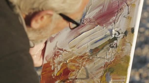 Senior man paints a picture on the beach. Over the shoulder close-up of elderly male artist in glasses applying paint to canvas with a spatula at pebble sunrise sea beach. — Stockvideo