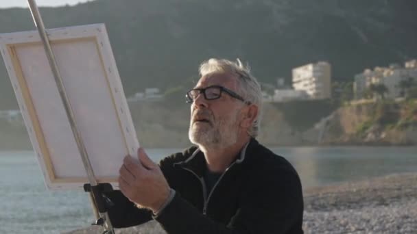 Un aîné peint un tableau sur la plage. Plan médian d'un artiste masculin âgé en verre peignant la toile au coucher du soleil plage de la mer . — Video