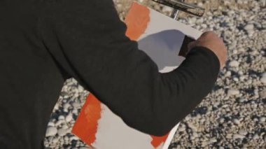 Komuta sizde boya bir resim sahilde. Çakıl gündoğumu deniz Plajı'nda geniş spatula ile tuvale akrilik boya uygulama yaşlı erkek sanatçı.