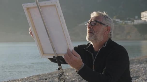 Starszy człowiek maluje obraz na plaży. Podeszłym wieku artysta w szkle, malowanie na sztalugach metalowe o zachód słońca sea plaża płótnie. — Wideo stockowe