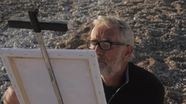 Ein älterer Mann malt ein Bild am Strand. Hochwinkelaufnahme eines älteren männlichen Künstlers mit Brille, der auf Leinwand auf einer Staffelei am Kieselstrand zeichnet. — Stockvideo