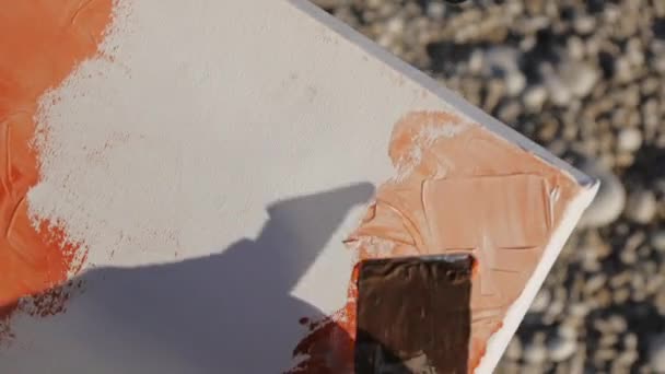 Senior man schetst een beeld op het strand. Over de schouder close-up shot van oudere mannelijke artiest acrylverf toe te passen op het canvas met brede spatel bij zonsopgang zee kiezelstrand. — Stockvideo