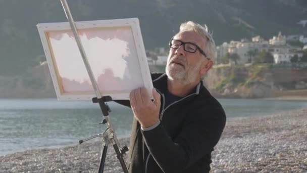 老人在海滩上画了一幅画 在日落海滩的金属画架上 在白色画布上涂上白色画布的老年男艺术家 — 图库视频影像