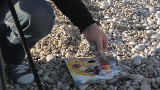 Senior man schetst een beeld op het strand. Oudere mannelijke artiest verf uit het palet te nemen en toe te passen op canvas met een spatel bij zonsopgang zee kiezelstrand. — Stockvideo