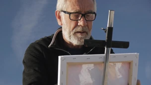 Senior man schildert een foto op het strand. Lage hoek uitzicht van oudere mannelijke kunstenaar schilderij van het doek met een brede spatel op Pebble Sunrise Sea Beach. — Stockvideo