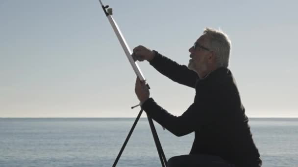 Ein älterer Mann malt ein Bild am Strand. ältere männliche Künstler Malerei der Leinwand auf Metall Staffelei am Morgen ruhige See Hintergrund. — Stockvideo