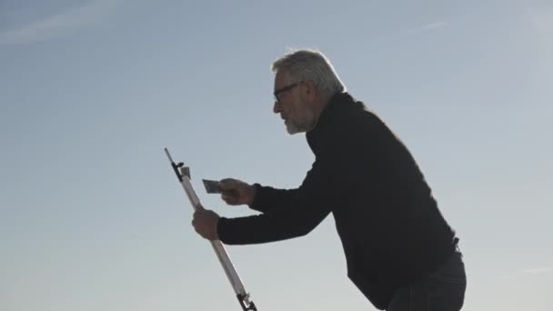 Un aîné peint un tableau sur la plage. Artiste masculin âgé peignant la toile sur chevalet métallique sur fond de ciel calme matinal. Plan moyen . — Video