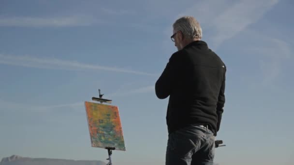 老人在海滩上画了一幅画。低角度背景的老年男性艺术家看他最近画的蓝天背景图片. — 图库视频影像