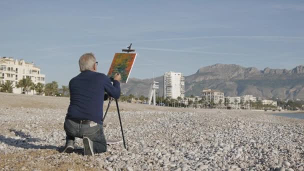 Un aîné peint un tableau sur la plage. Vue arrière d'un artiste masculin âgé peignant une image abstraite avec un pinceau sur fond de ville balnéaire . — Video